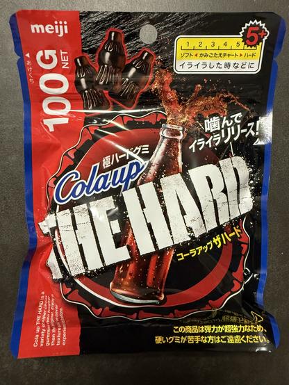 A colorful packaging for a candy product called "Colaup THE HARD" by Meiji. The package features a graphic of soda and includes a warning about the candy's hardness. It displays the net weight of 100g and indicates that it can be