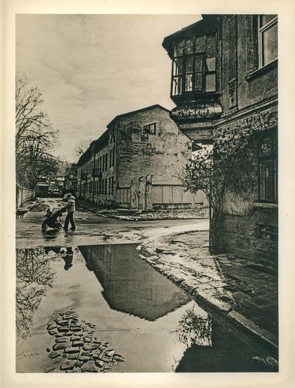 This is a black-and-white photograph created using the lith print technique in a darkroom. The image has a characteristic high-contrast and grainy texture, with deep blacks and soft highlights, typical of this alternative photographic process.

The scene depicts an old, weathered urban street with aged buildings that show signs of wear and decay. On the right, there is a corner of a historic-looking building with intricate detailing and a protruding bay window. The background features additional old structures, some with cracked walls and peeling plaster.

A large puddle in the foreground reflects the surrounding buildings, trees, and sky, adding depth and symmetry to the composition. The reflection creates an almost surreal effect, making the ground appear as a second sky.

In the middle of the street, a woman is pushing a baby stroller, adding a human element to the scene. The mood of the image is nostalgic and atmospheric, enhanced by the lith print technique, which gives it a timeless and slightly eerie quality.