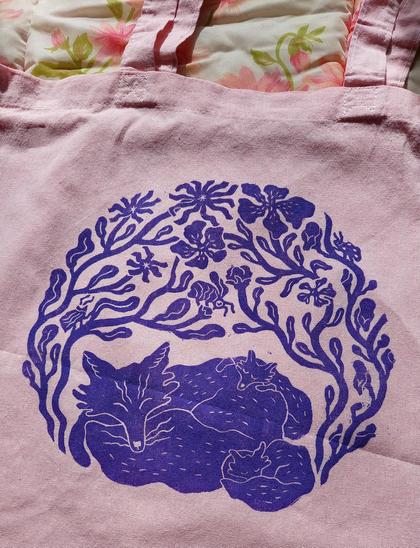 A purple linocut print on light pink fabric. The print shows three foxes huddled together and looking happy, hidden under a canopy of plants and flowers. Three insects are hidden inside the plants and kind of watching over the foxes. There are a bee, a beetle and a ladybug.