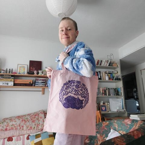Flore (the artist) holding a pink tote bag printed with a purple ink linocut. (Same print than the first picture) Flore is smiling.
