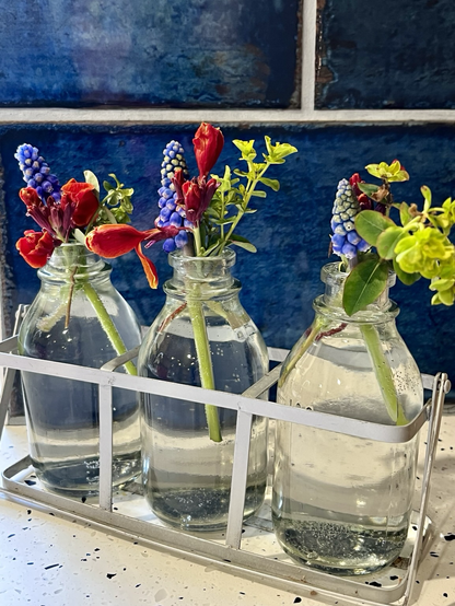 Small milk bottle vases with green euphorbia, orange wallflowers and blue muscari. Dark blue tiles on wall behind.