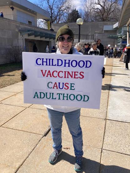 A smiling person wearing sunglasses, a green beanie, and casual clothing holds a humorous protest sign outdoors that reads, 'CHILDHOOD VACCINES CAUSE ADULTHOOD,' parodying typical anti-vaccine slogans. The scene takes place in a sunny area with several people visible in the background, suggesting a gathering or rally.