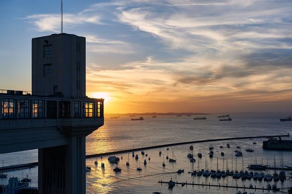 The photo shows a sunset by the sea. On the left an historical elevator building is covering the sun. On the font right sailing boats in the port are seen. The upper side of the sky shows the blue sky with stripes of clouds. Towards the horizon the colors shift from blue to orange.