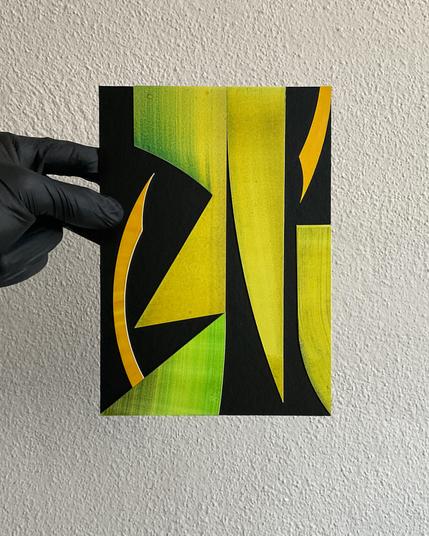 A5 collage-art held in a gloved hand on a cream textured wall. The collage shows yellow and lime green geometric paper-cutout shapes on a black background.