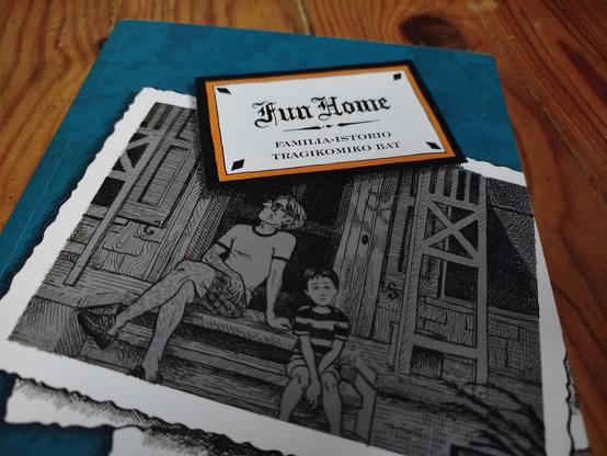 Fun Home komikia egurrezko mahai baten gainean. Fondo berdean argazki familiar bat, aita eta alaba bat etxe atarian.