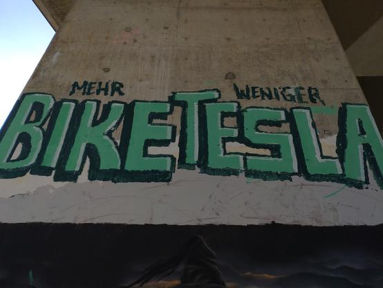 Ein Graffito an einem Betonpfeiler einer Autobrücke angebracht. In kleinen Buchstaben steht dort Mehr darunter in riesigen grünen Buchstaben das Wort Bike. Rechts daneben in kleinen Buchstaben weniger und in riesigen das Wort Tesla. Zusammen ergibt das: Mehr Bike weniger Tesla.