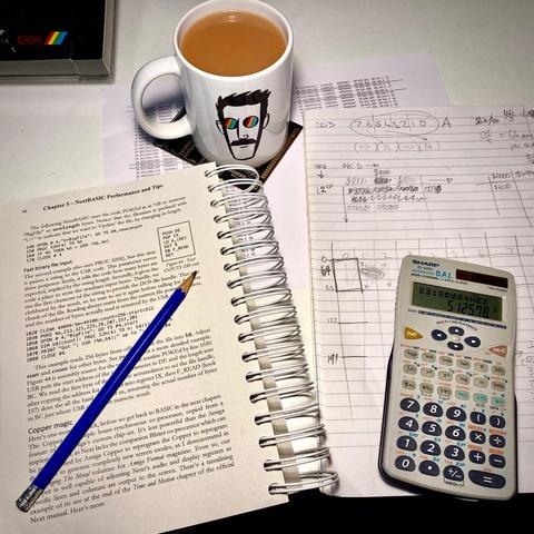 A photograph of my desk with a book, a notepad, a pencil, a mug of tea and a calculator.