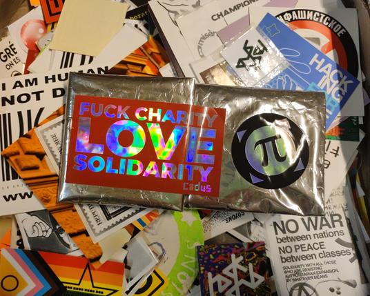 le premier prototype de pochette dont la surface brillante en alu et partiellement recouverte de stickers sur papier métallisé irisant. à gauche, celui de l'ONG cardus écrit "fuck charity, love solidarity". à droite, le logo de la quadrature du net. la pochette est posée sur un tas de stickers wokistes.