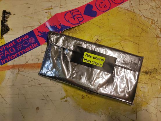 le premier prototype de pochette dont la surface brillante en alu et partiellement recouverte de stickers sur papier métallisé irisant. à gauche, celui de l'ONG cardus écrit "fuck charity, love solidarity". à droite, le logo de la quadrature du net. la pochette est posée sur un tas de stickers wokistes.