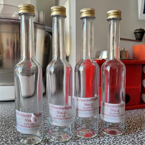 Foto von vier kleinen, durchnummerierten Probierflaschen mit der Aufschrift "Krisenwhisky". Im Hintergrund eine Küchenmaschine und eine manuelle Kaffeemühle.
