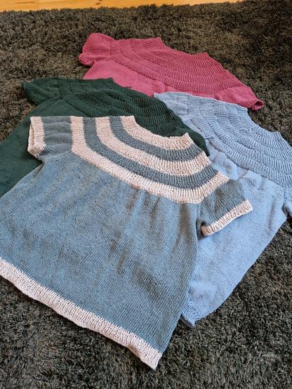 Fyra stycken stickade kortärmade sommartröjor i olika färger ligger utlagda lite omlott på en fluffig matta.