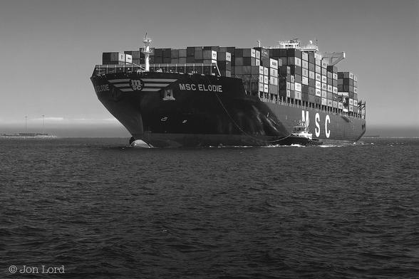 A Black And White Seascape Photo In Landscape Format. From The Base Of The Image Upwards To The Halfway Point Is A Body Of Black Smooth Seawater. Slightly Below The Horizon Is A Large Container Ship, Filling About Two-Thirds Of The Photo, Underway From Right To Left. The Ship Has A Black Hull And White Superstructure. On The Upper Part Of The Bow Are Three White Chevrons With The Letters Of The Shipping Line 'MSC' In The Centre. Aft Of The Chevrons Again In White Is The Ships Name 'MSA ELODIE'. On The Ship's Deck Are Stacks Of Shipping Containers, Seven High In Places. About One Quarter Of The Way Aft Of The Bow Is A Large Tug, Dwarfed By The Ship, And Attached With A Slack Line That's Secured To The Bow. The Ships Bridge And Accommodation Superstructure Located Forward Of The Stern Is Just Visible Above The Stacked Containers. To The Left Of Our Ship A Few km Away Is A Short Breakwater With Two Tall Floodlights. In The Distance Is A Horizon With A Thin Layer Of Dark  Haze. Above: A Clear And Cloudless Sky. 
The Location: The Port Of Long Beach, California (2016).