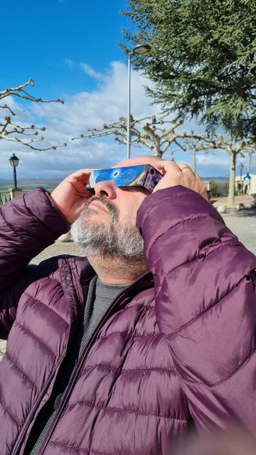 Estoy mirando hacia el sol con unas gafas de eclipse homologadas. Llevo un plumífero de robo berenjena. Me sujeto la gafas con las manos. Al fondo un parque con árboles.