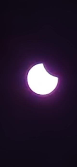 Stark gezoomtes und dadurch leicht unscharf wirkendes Bild der partiellen Sonnenfinsternis auf dem Höhepunkt. Der Mond deckt oben rechts die Sonne ab. Durch den als Filter verwendeten schwarzen Diafilm erscheint das Bild leicht violett eingefärbt.