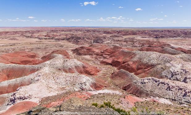 Looking down into a wide basin with eroded hills of colorful soils: reds, whites, orange, and brown; little vegetation; blue sky with a few small clouds along the horizon.
©BosqueBill.com