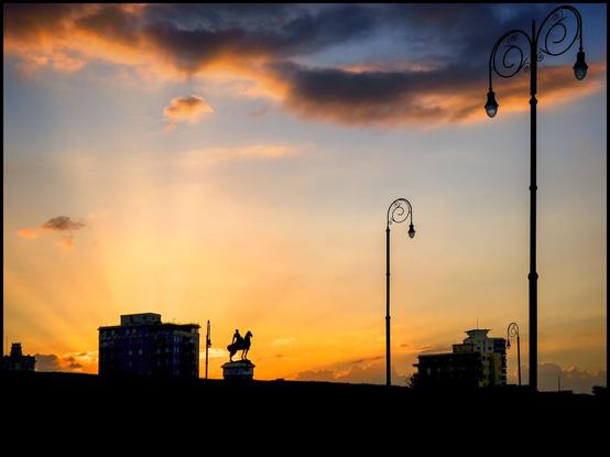 "A sunset photograph showing a silhouette of a city skyline with multiple buildings, several ornate street lamps, and a statue of a person on horseback. The sky is filled with warm tones of orange and yellow, indicating dusk or sunset."