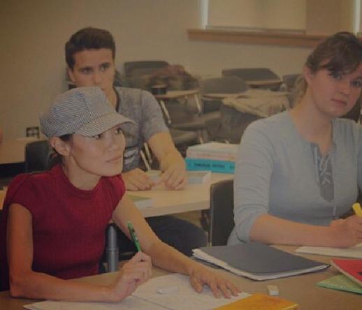 Three students sitting at desks in a classroom. Me in the foreground, wearing a newsboy cap and red shirt, staring ahead while holding a green pen. A male student sits behind her to the left, and a woman with brown hair sits to the right in a blue shirt. They're all writing in notebooks or on paper. Textbooks and other school items are scattered on the desks. An empty row of desks is in the background.