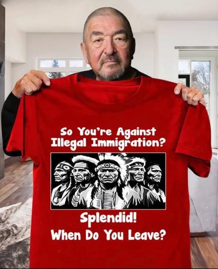 First Nations actor, Graham Green, standing in a black shirt Holding up a large red T-shirt with a photo Of five first nation Chiefs. Above in white lettering.” So you’re against illegal immigration? Below the Photo “ Splendid, when do you leave?”