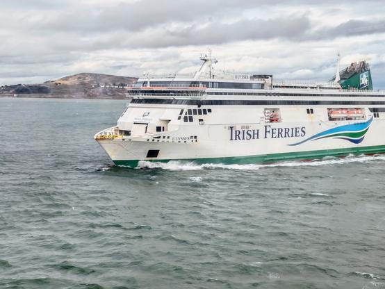 Picture of marine vessel RO-RO ferry Ulysses operated by Irish Ferries in the navigation channel leading into Dublin port. The bow faces to the left of frame. The peninsula of Howth is visible behind in the background.