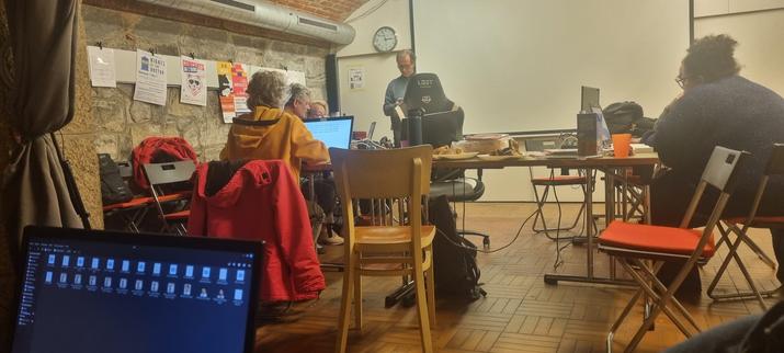 Photo du local de la SDMB avec des tables placées en carré avec diverses personnes autours et du matériel informatique.