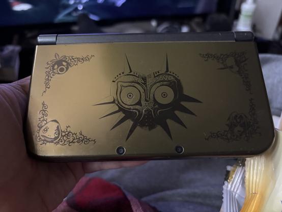 A person is holding a limited edition gold-colored Nintendo 3DS XL featuring a black etched design from The Legend of Zelda: Majora’s Mask. The central artwork showcases Majora’s Mask, with intricate corner decorations depicting characters and masks from the game. The background includes a dimly lit room with a television screen and various household items.