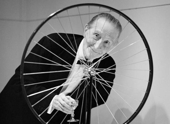 Portrait de Marcel Duchamp, tenant un verre, et posté derrière une roue de biciclette.
