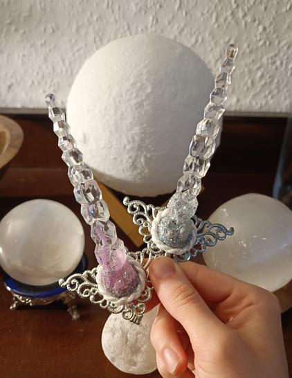 A hand is holding two unicorn horns. One of them has some pink glitter at the bottom and one some blue. The horns are clear and look crystally. In the background are standing four white spheres.