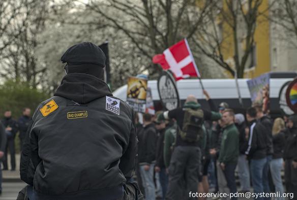 Eine Person  mit aufnäher auf dem rücken ("skins gegen nazis", "bad example") sitzt neben dem demozug und bepöbelt diesen