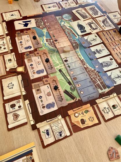 A tabletop game setup featuring a detailed game board with a map design and various cards placed around it. Player markers and tokens are visible on the board, along with several action and item cards. The game components include illustrations related to ships and resources, with
