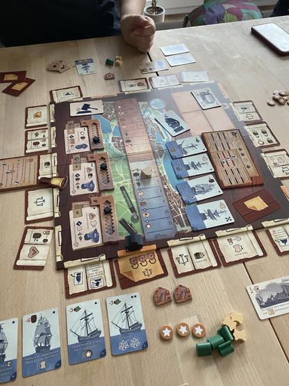 A tabletop game in progress on a wooden surface. The game board features various cards, tokens, and player pieces, including ships and resources. A person's hand is visible, interacting with the game components. The overall setting appears casual and inviting.
