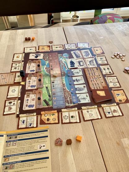 A tabletop game setup featuring a board with various cards, tokens, and player pieces, including wooden cubes and a highlighted instruction sheet. The game appears to involve resource management or strategy, with a focus on building or trading elements. The scene is set on
