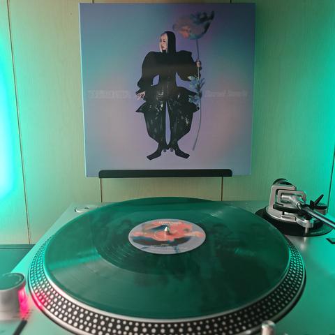 A Winter Green vinyl record sits on a turntable. Behind the turntable, a vinyl album outer sleeve is displayed. The front cover shows TOKiMONSTA wearing hooded black outfit that is bulging out at the bottoms. She is holding a large flower.