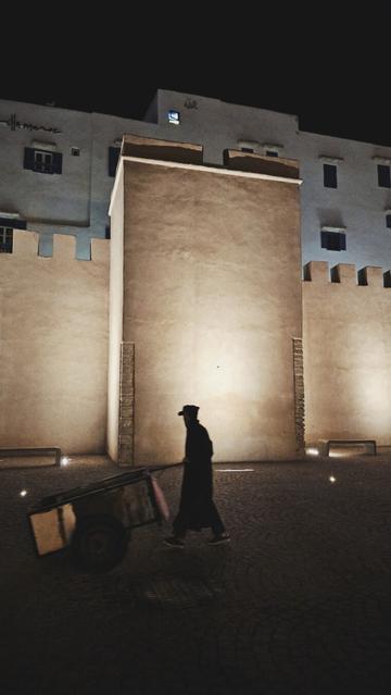 The portrait-orientated image shows a person pushing a two-wheeled cart on a paved surface at night. The person is silhouetted, wearing a long robe and a cap. Behind the person and cart are several light-colored, rectangular structures with crenellated tops, belonging to the city walls and buildings. The walls is illuminated by artificial lighting, from the ground, bringing out the texture of the yellowish city walls.