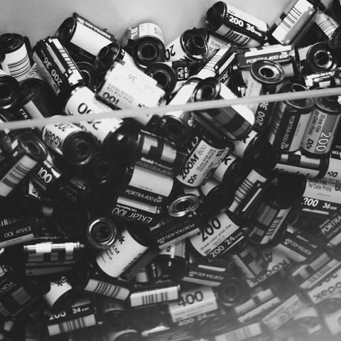 A monochrome photo showing a large collection of 35mm film canisters, including Kodak Portra 400, Kodak Ektar 100, and Kodak Zoom 200. The canisters are densely packed in a container, creating a visually textured and nostalgic composition.