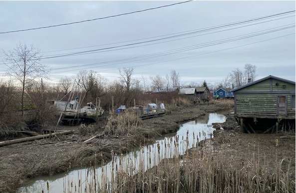 Image of a creek flowing through Finn Slough, near Richmond British Columbia.  Old buildings, bull rushes, semi-abandoned boats, all under a grey sky.