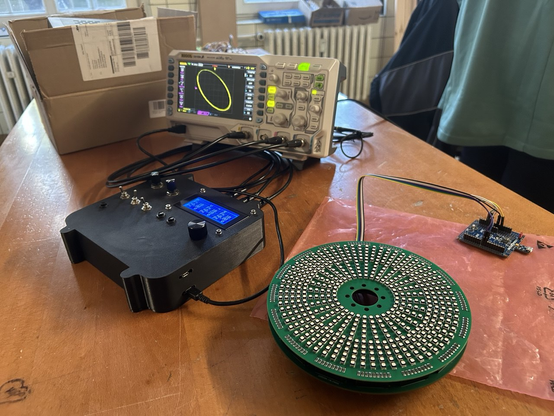 Tisch mitten im Raum mit Ozi, welches im XY-Modus eine Ellipse zeichnet. Gespeist wird er aus einem Kasten mit schwarzem 3D-Gehäuse, diversen Kippschaltern und Potis. Daneben ein rundes PCB um vielen Kreisen mit RGB-LEDs.