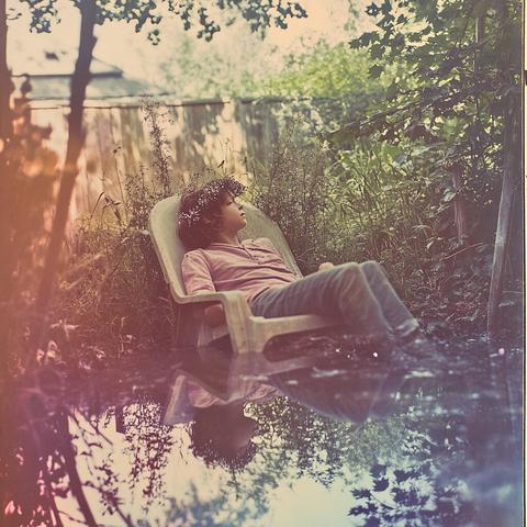 A dreamy, vintage-style image of a child lounging in a plastic chair in a lush backyard. They wear a pink shirt, jeans, and a flower crown. The ground is wet, reflecting their image like a mirror, enhancing the ethereal, fairy-tale vibe.
