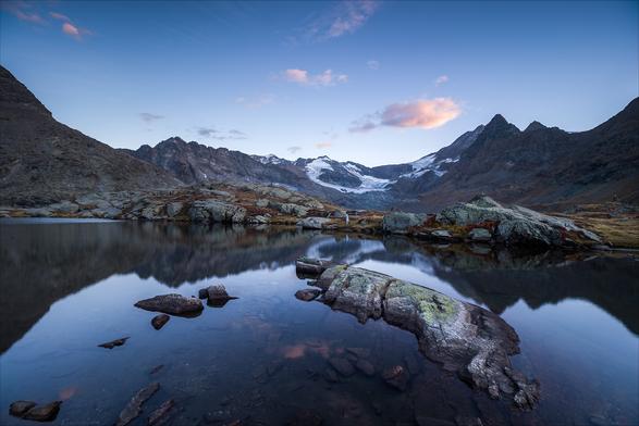 Photo d'un paysage de montagne au coucher du soleil.
Au premier des cailloux baignent dans un petit lac. L'un deux, beaucoup plus gros et aplati, pourrait évoquer une forme d'animal couché ou de cétacé échoué.
En arrière plan l'horizon est fermé par de hautes montagnes, on peut voir des glaciers qui en descendent.
Dans le ciel bleu passent quelques petits nuages roses.
-
Photo of a mountain landscape at sunset.
In the foreground, pebbles bathe in a small lake. One of them, much larger and flattened, could evoke the shape of a recumbent animal or beached cetacean.
In the background, the horizon is closed in by high mountains, from which glaciers can be seen descending.
A few pink clouds pass through the blue sky.