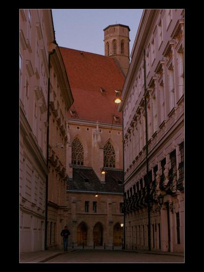 Color photograph showing the view of a Viennese alleyway, taken shortly before dusk. The Minoritenkirche church can be seen at the end of the alley. On the sidewalk to the right, a person is walking towards the camera, looking down at the watch on their wrist.
