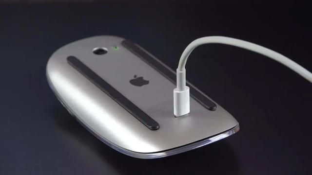 La imagen muestra la parte inferior de un Apple Magic Mouse, donde se puede ver el puerto de carga. Un cable blanco está conectado al puerto. El Magic Mouse tiene un acabado plateado con detalles negros en los patines deslizantes. Se aprecia el logo de Apple. El fondo es negro, lo que ayuda a resaltar el diseño del ratón.