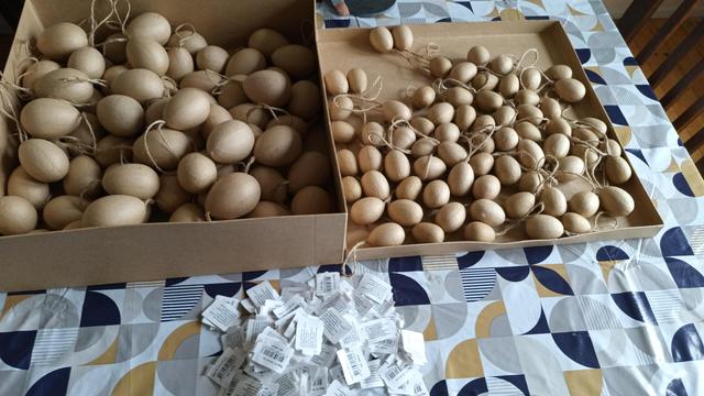 Large square cardboard box containing 70 large brown paper mache eggs, the lid is next to it with 70 small eggs 
There is a pile of labels (removed from the eggs in front of the boxes