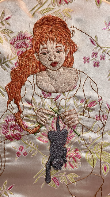 An embroidered picture of a red haired woman knitting a kitten.