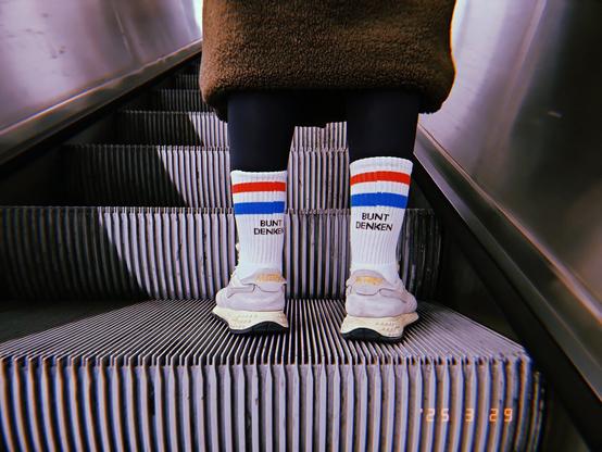 A person is standing on an escalator's stair with only the tennis socks in the focus. The German words "bunt denken" are written on the socks.