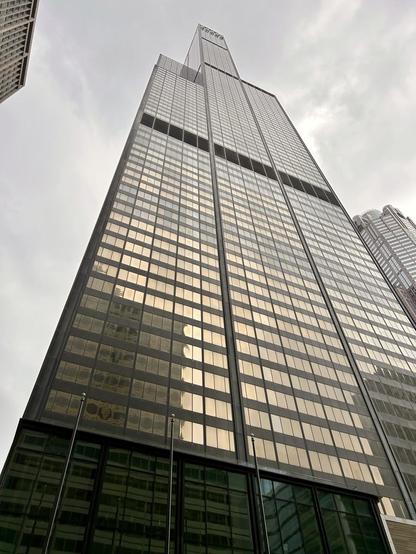 A low-angle picture of the Sears Tower (it’s still called that shut up), its tinted windows reflecting a golden-bronze color against a cloudy sky.