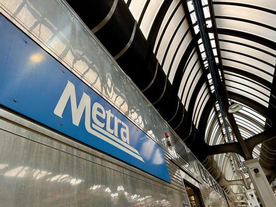 A low-angle picture of a silver bi-level train car emblazoned with “Metra” in white on a blue panel. Daylight shines in diffusely through the frosted glass canopy above.