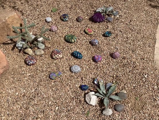 Around fifteen small rocks with colorful crochet covers, in a gravel yard among agave and cholla.