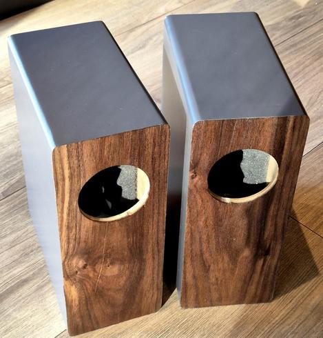 A pair of speaker boxes on a grey wooden floor. The boxes are grey, wrapped in vinyl, with walnut veneer fronts. The drivers are still missing, I haven't put them in yet.