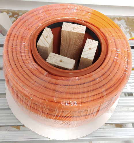 DIY cable reel with the shrink-wrapped cable placed on it. One side wall is still missing so the interior construction is visible.
Four pieces of wood connect the side walls together. They are surrounded by a pipe sleeve, providing a smooth surface for the cable to rest on. There's only a minimal gap between sleeve and cable.