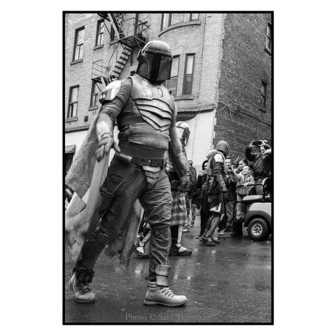 (English Follows). Photographie noir et blanc, format portrait, ratio 2x3. Un cosplayer porte un costume de Mandalorian. Sa "cape" est un drapeau irlandais. La photographie est prise en contre-plongée. Le Mandalorian avance, fixant l'objectif. À l'arrière-plan, se distinguent un autre Mandalorian en interaction avec le public, ainsi qu'un bâtiment typiquement montréalais (briques et escalier de secours en métal). 

Black and white photograph, portrait format, 2x3 ratio. A cosplayer wears a Mandalorian costume. His “cape” is an Irish flag. The photograph is taken from a low angle. The Mandalorian moves forward, staring into the lens. In the background, another Mandalorian can be seen interacting with the audience, as well as a typical Montreal building (bricks and metal fire escape).