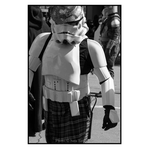 (English follows) Photographie noir et blanc, format portrait, ratio 2x3. Un Stormtrooper porte un kilt, et un chapeau avec plusieurs shamrocks. La photographie est prise de face, le Stormtrooper occupe l'essentiel de l'image. À l'arrière plan, on distingue un Mandalorian. 
Black and white photograph, portrait format, 2x3 ratio. A Stormtrooper wears a kilt and a hat with several shamrocks. The photograph is taken from the front, with the Stormtrooper occupying most of the image. A Mandalorian can be seen in the background.